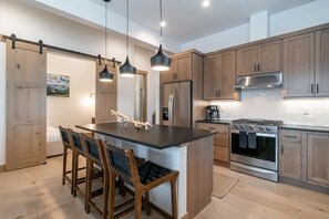 Una cucina moderna con mobili in legno, un'isola centrale, illuminazione a sospensione, un frigorifero, un forno e un'area pranzo con sedie.