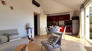 Una sala moderna con sofá, una mesa de centro y un comedor con sillas y mesa. Se aprecia una cocina al fondo con armarios rojos y un refrigerador.