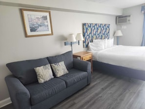 Habitación de hotel con sofá gris, cama, mesita de noche, lámpara, aire acondicionado de pared y un cuadro enmarcado.