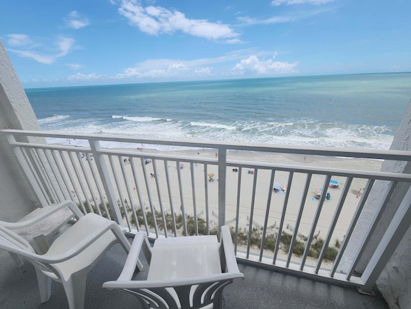 A balcony with white plastic chairs, a view of the beach, and the ocean.