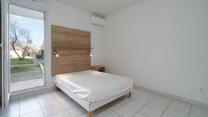 A bedroom with a wooden headboard, a bed with white bedding, a bedside table, and a view through an open door to a garden.