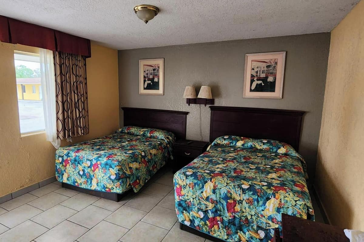 Quarto com duas camas, janela com cortinas, luminária fixada na parede e quadros emoldurados.