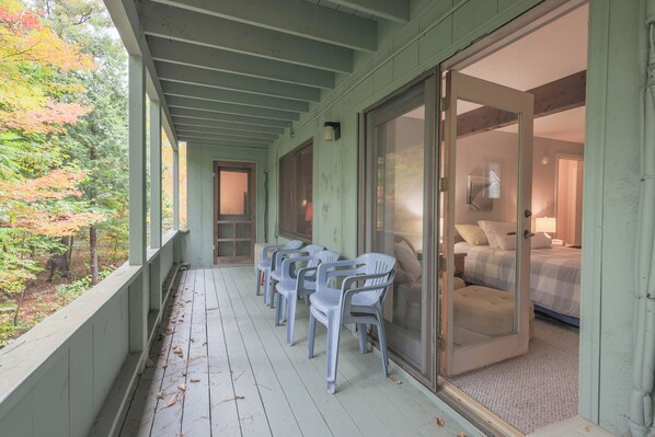 A covered porch with plastic chairs, a sliding glass door to a bedroom, and a view of trees.