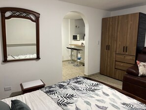 Relax in a serene bedroom with a large bed and a stylish wardrobe, featuring a kitchenette area with a microwave and bar stool.