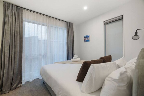 A hotel room with a bed, curtains, a wall-mounted lamp, and a window with sheer curtains.