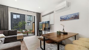 Un salon moderne avec un canapé, une table à manger et un téléviseur. Il y a une fenêtre avec des rideaux, une plante et un tableau au mur.
