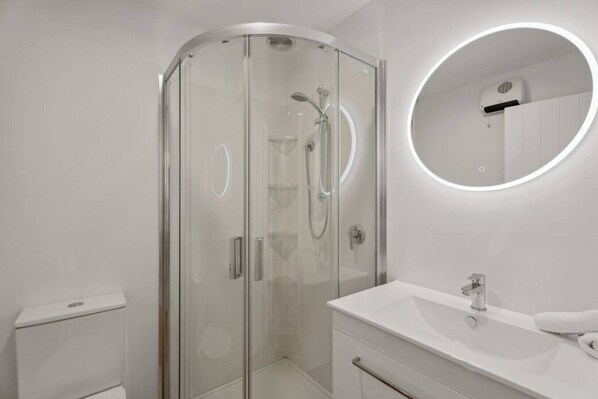 Un baño moderno con cabina de ducha de vidrio, un lavabo blanco y un espejo redondo con iluminación integrada.