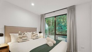 Ein modernes Schlafzimmer mit einem großen Bett, einem Nachttisch, einer Lampe und Blick auf einen Balkon mit Vorhängen.