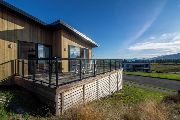A modern wooden house with a deck, large windows, and a view of a road and distant mountains.
