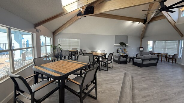 Een ruime loft-achtige kamer met een hoge plafond, zichtbare houten balken, een eethoek met een houten tafel en stoelen, een zithoek met banken en een salontafel, en grote ramen met uitzicht op buiten.