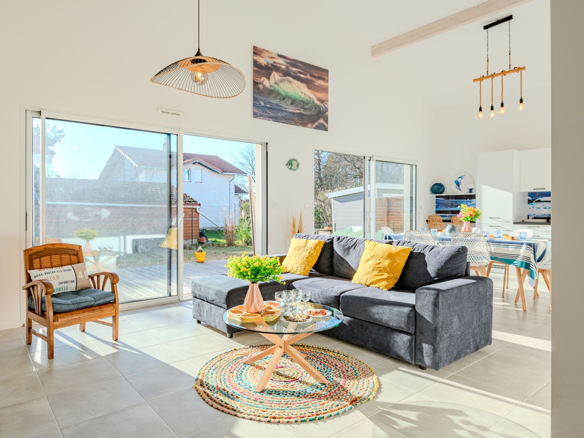 Een moderne woonkamer met een grijze bank, een houten stoel en een glazen salontafel. Er ligt een kleurrijk geweven vloerkleed, een hanglamp en een schilderij aan de muur.