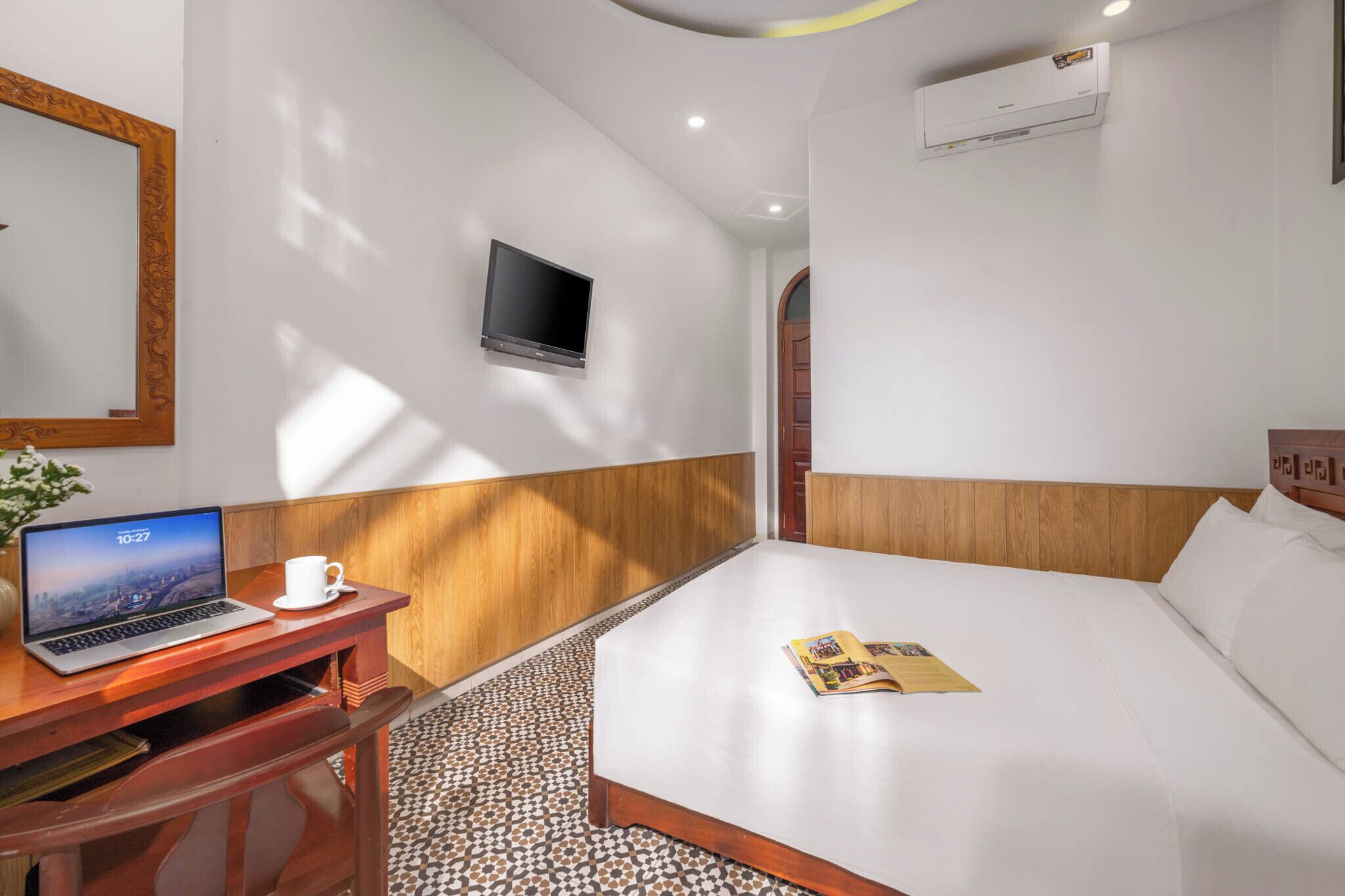 A hotel room with a bed, a desk with a laptop, a TV, and an air conditioning unit.