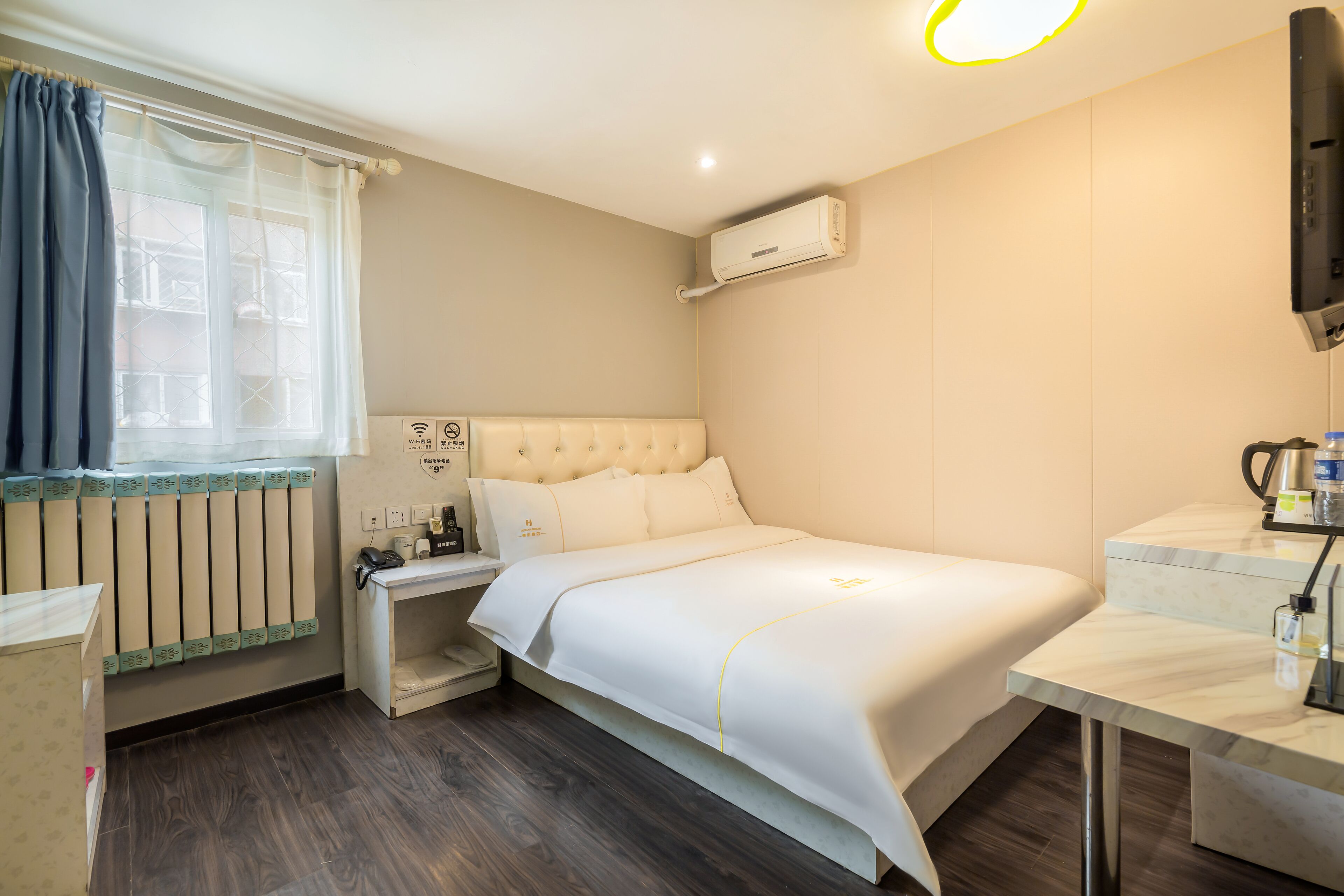 A hotel room with a bed, a bedside table, a nightstand, a window with curtains, a radiator, an air conditioner, and a television.