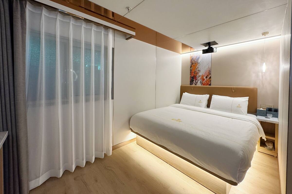 A modern hotel room with a large bed, a wooden headboard, a nightstand, and a large window with sheer curtains.