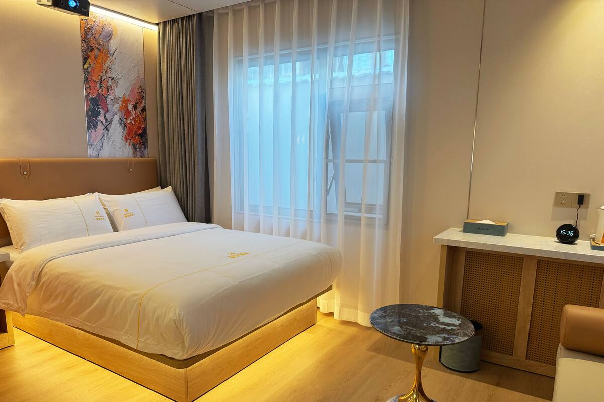 A hotel room with a large bed, a bedside table with a clock, a small round table, and a window with sheer curtains.