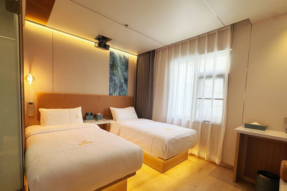 A hotel room with two beds, a desk, a small table, a projector, and a window with curtains.