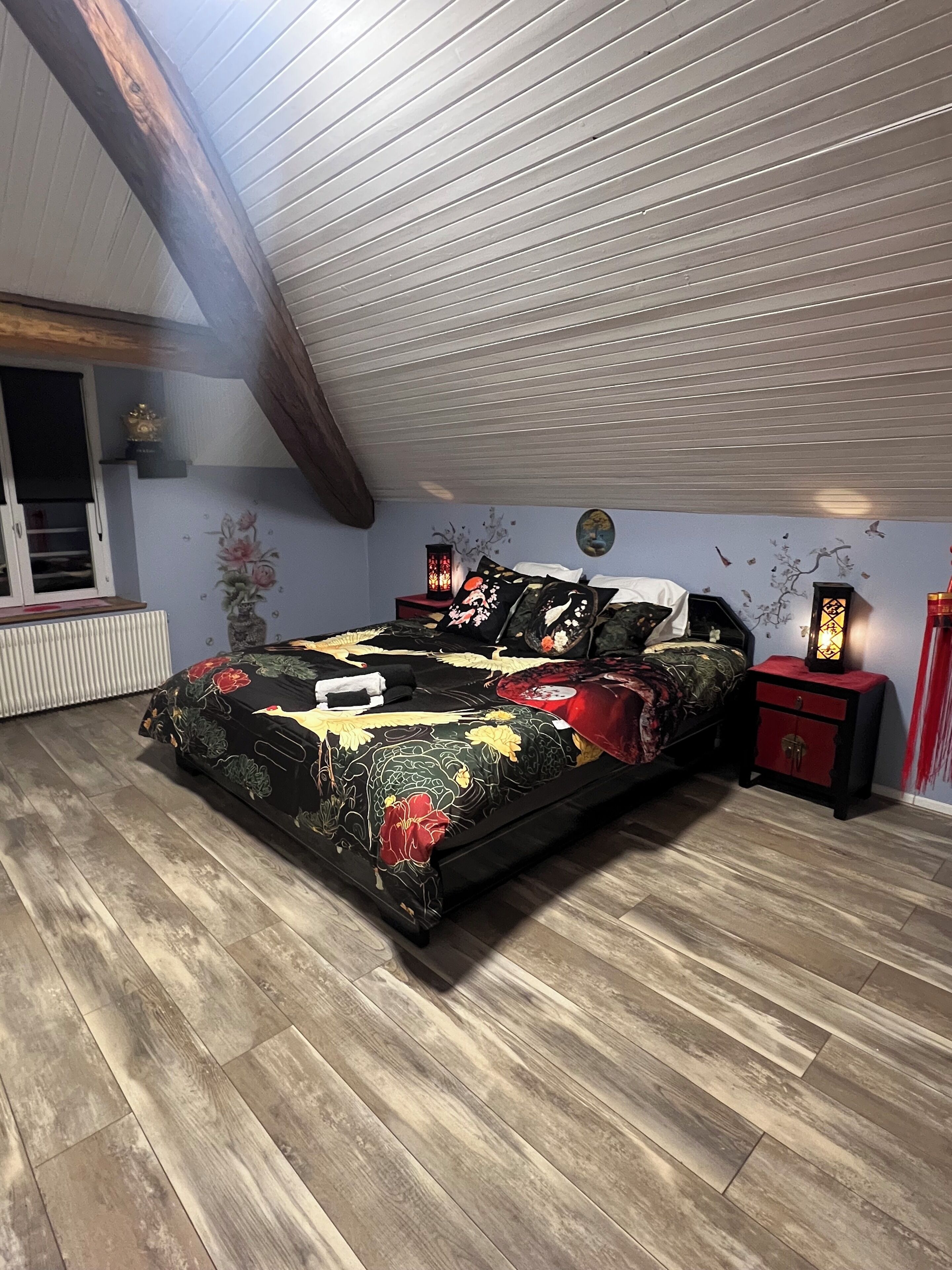 Une chambre avec un plafond en pente, un lit recouvert d’une literie à motifs floraux, deux tables de chevet avec des lampes et un radiateur.