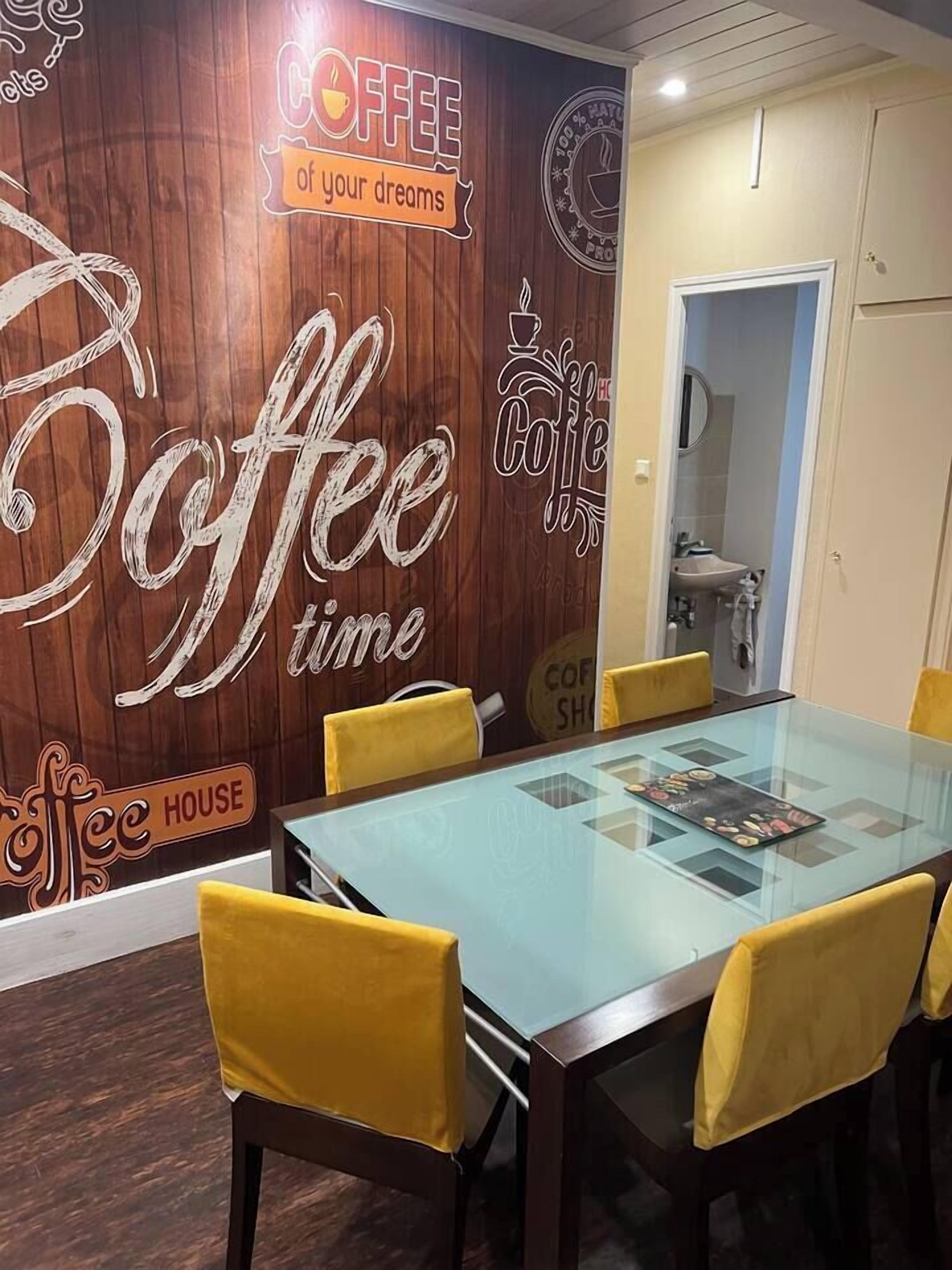 A meeting room with a glass table and yellow chairs, a wooden wall with coffee-themed decorations, and a visible bathroom door.