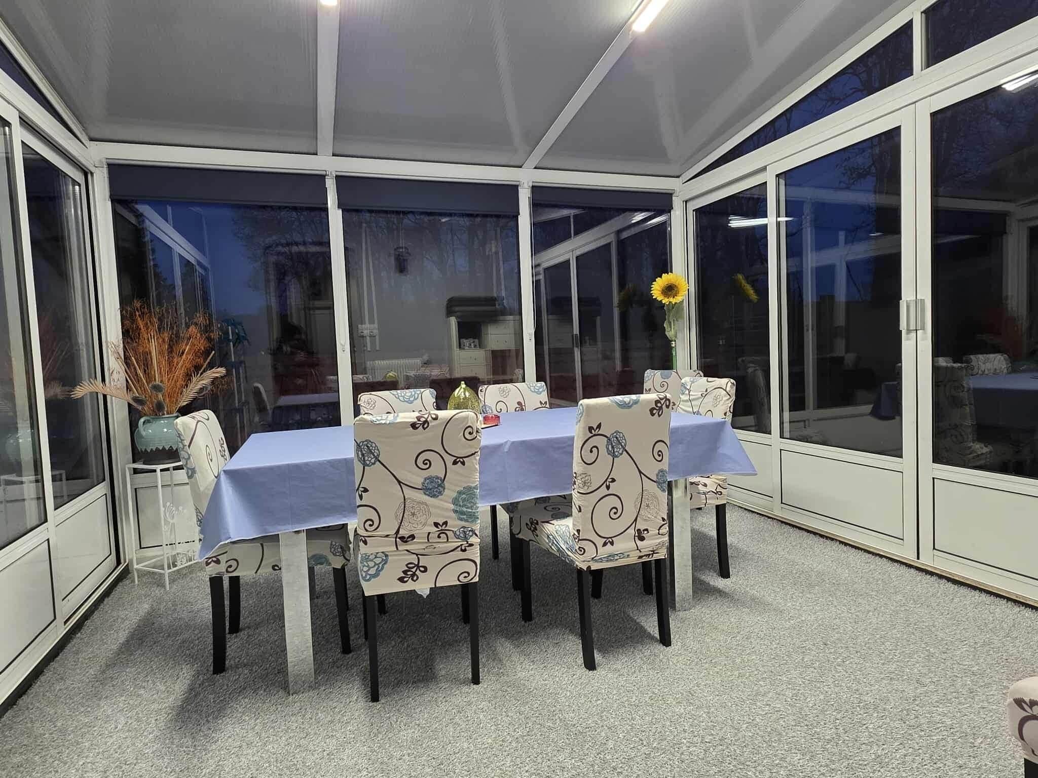 Eine überdachte Terrasse mit Tisch und Stühlen, großen Fenstern und einer Vase mit einer Sonnenblume.