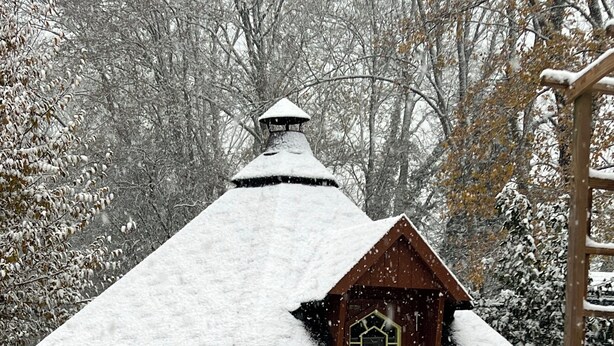 Una pequeña cabaña de madera cubierta de nieve con techo inclinado, ventana hexagonal y un porche nevado.