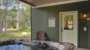 Un bain à remous avec une chaise en bois placée devant, un panneau d’avertissement et une porte dotée d’une fenêtre donnant sur des arbres et une maison.