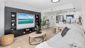A modern living room with a flat-screen TV, a sofa, a coffee table, and a kitchen area with a dining set. - #2 Dreamscapes Oceanfront Oasis, Sea Pointe 605 (North Myrtle Beach)