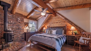 A cozy attic bedroom with a wooden ceiling, a brick fireplace, a bed with a plaid bedspread, and a nightstand with a lamp.