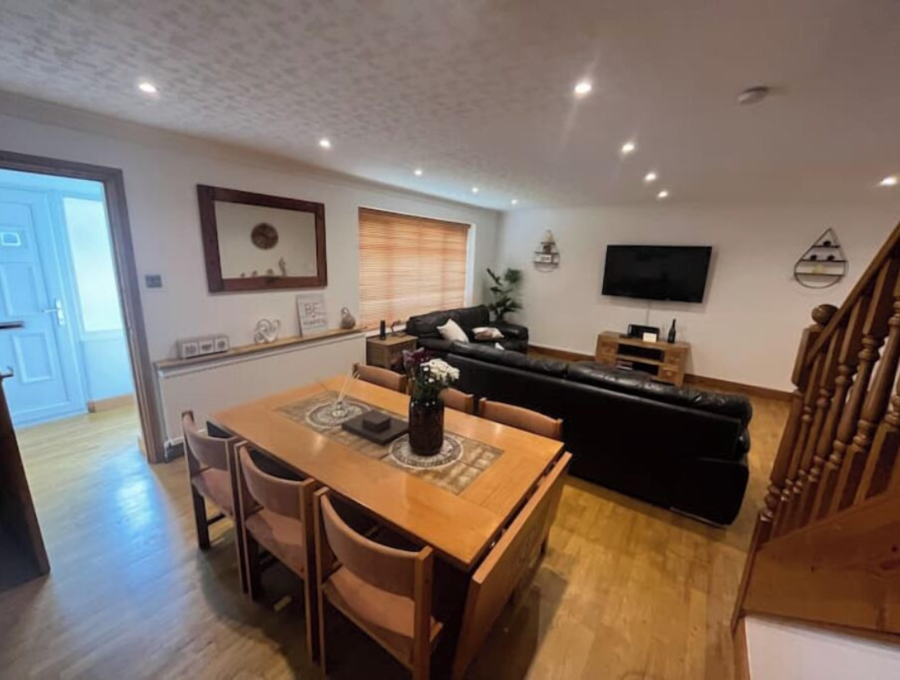 Een woonkamer met een zwarte leren bank, een houten eettafel en stoelen. Er is een televisie aan de muur bevestigd en een trap aan de rechterkant.