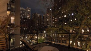A rooftop terrace with a view of a cityscape at night, featuring a wooden deck, outdoor seating, and a glass balustrade.