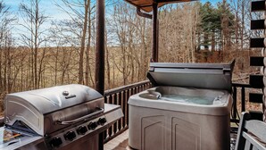 An outdoor grill and hot tub setup on a wooden deck with a view of a forested area. - "Ultimate Luxe Escape with Private Movie Theater and Game Room" (Nelsonville)