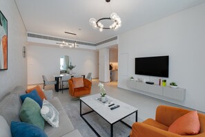 Un salón moderno con sofá, mesa de centro y sillón naranja. Un televisor de pantalla plana está montado en la pared, y se observa un área de comedor al fondo.