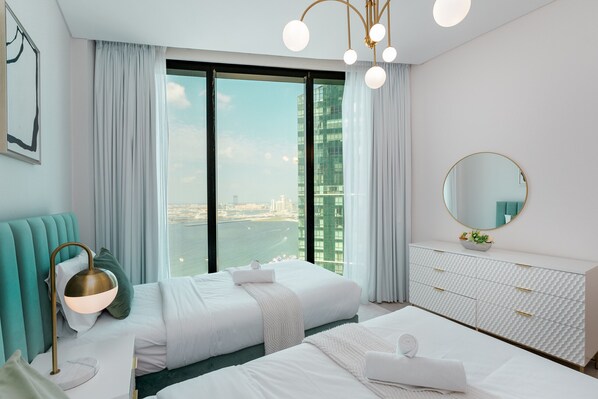 A modern hotel room with a large window, a bed, a bedside table, a lamp, a dresser, and a mirror.