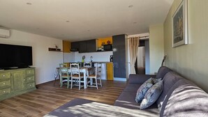 A living room with a television, a green cabinet, a dining area with chairs, and a kitchen in the background. - Mini villa T3 air-conditioned, sea view, cosy & modern (Olmeto)