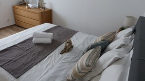 Una cama bien hecha con un plaid gris, ropa de cama blanca y almohadones decorativos. Una cómoda de madera con un espejo y una planta pequeña está al lado de la cama.