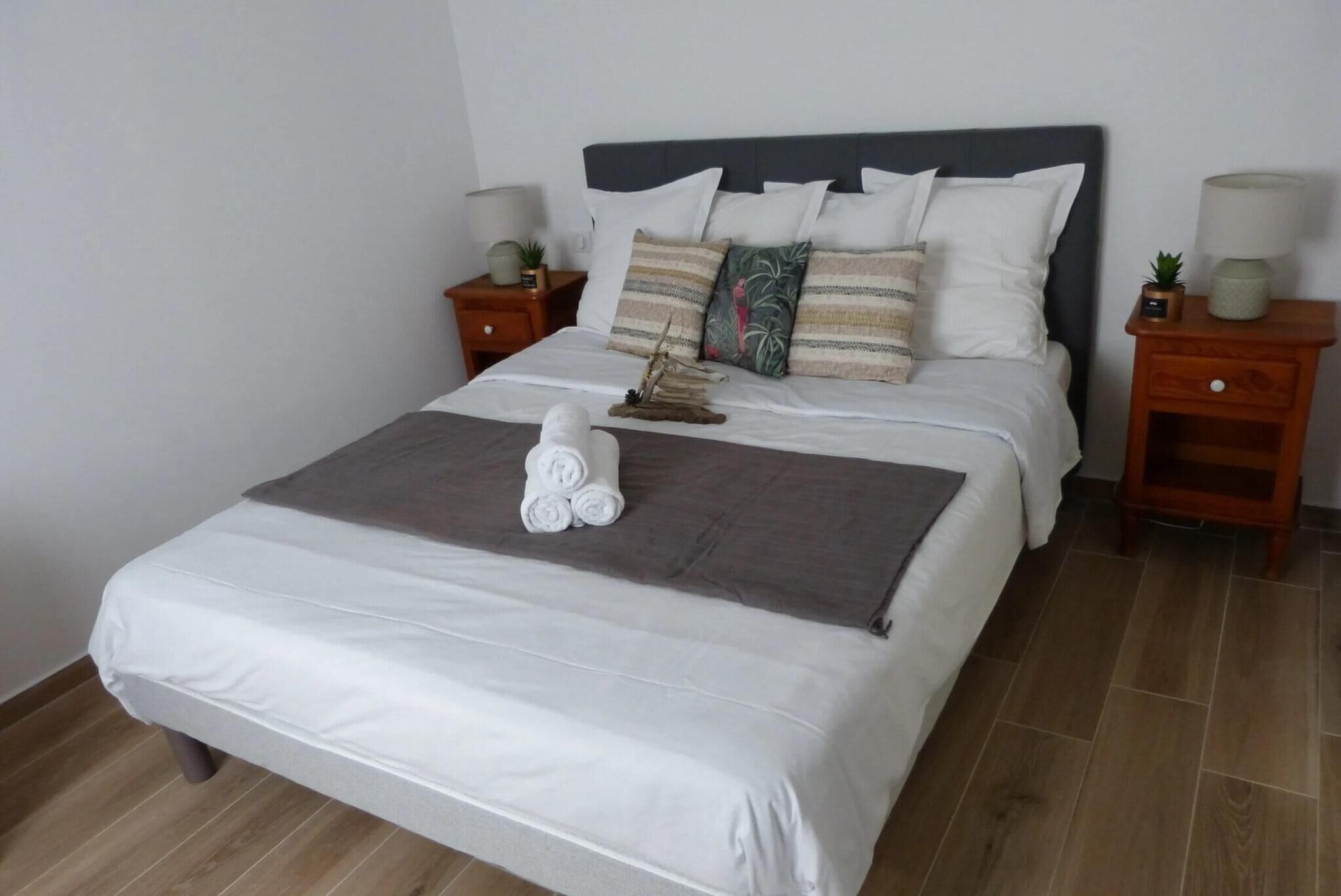 A neatly made bed with white and gray linens, a dark headboard, and decorative pillows. There are two bedside tables with lamps and small plants.