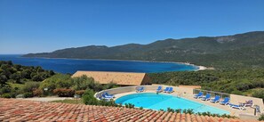 A swimming pool with blue chairs, a tiled roof, and a view of the sea and hills.