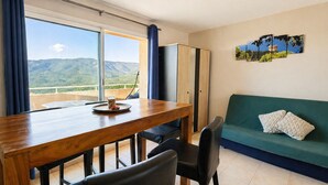 Una habitación con una mesa de comedor de madera, sillas negras, un sofá verde con un cojín estampado y vistas a montañas a través de un ventanal grande.