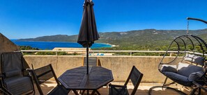 A terrace with a table and chairs, a hanging chair, and an umbrella, overlooking a coastal landscape with mountains.