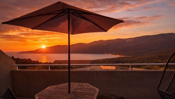 Un balcón con mesa y sillas, con vistas a la puesta de sol sobre un cuerpo de agua y montañas.