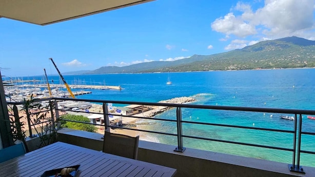 Un balcon avec vue sur une marina, une mer d’un bleu limpide et une montagne en arrière-plan.