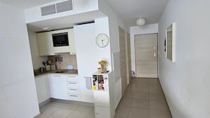 Une cuisine compacte avec des meubles blancs, un four à micro-ondes encastré et un petit plan de travail. Il y a une horloge au mur et une porte qui donne sur une autre pièce.