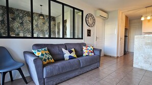 A living room with a grey sofa, a side chair, a large window, and a kitchen area with a brick backsplash.