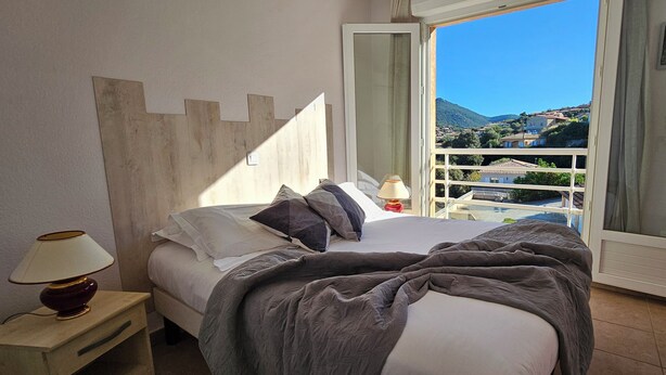 A bedroom with a bed, a nightstand, a lamp, and a view of a balcony with a cityscape.