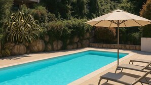 A rectangular swimming pool with clear blue water, surrounded by a tiled deck. There are lounge chairs and a single umbrella providing shade.