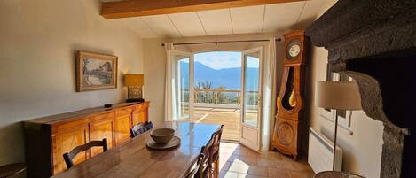 A dining area with a wooden table, chairs, a bowl, a cabinet, a clock, and a view of mountains through an open door.