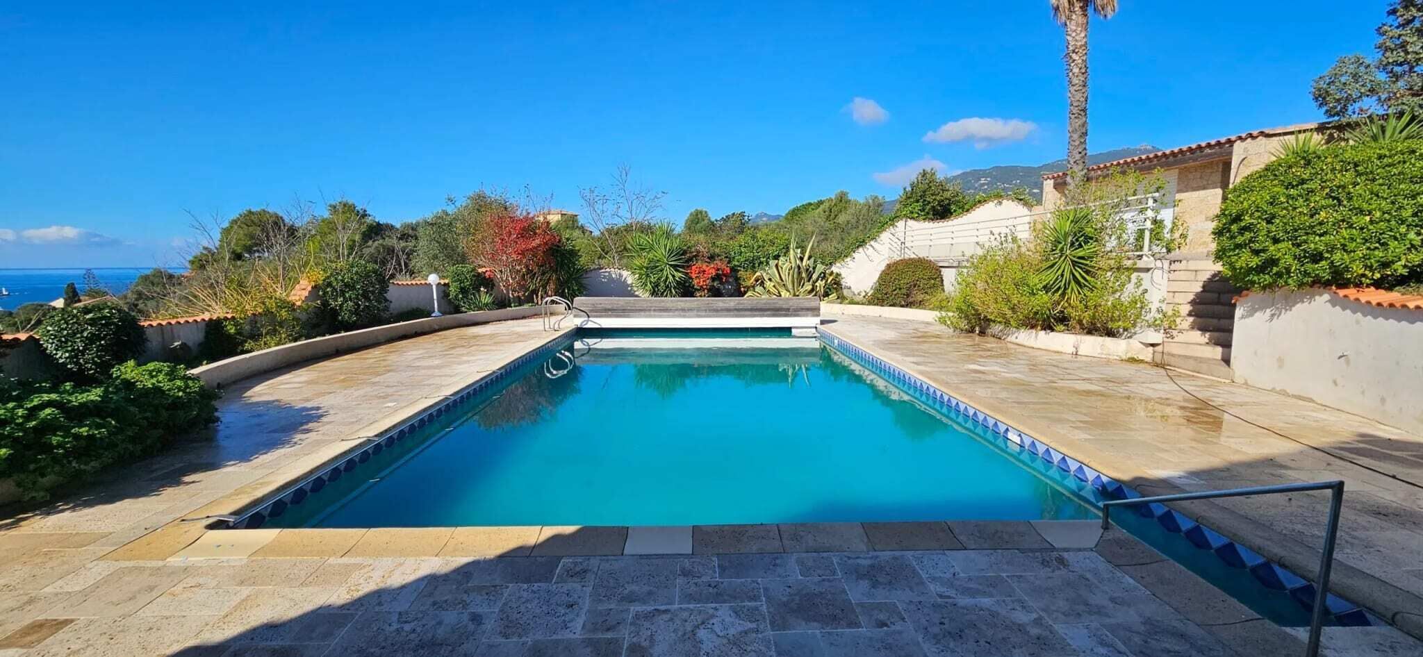 Ein rechteckiger Swimmingpool mit glasklar blauem Wasser, umgeben von Steinplatten und begrenzt durch eine niedrige Mauer.