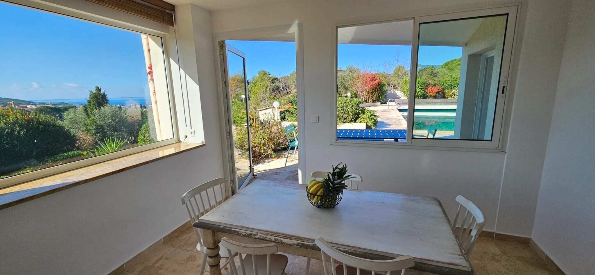 Ein Zimmer mit Tisch und Stühlen, einem Fenster mit Blick auf Grünflächen und einen Pool, sowie einer Tür, die nach draußen führt.