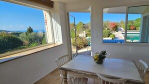 A room with a table and chairs, a window with a view of greenery and a pool, and a door leading outside. - Luxury villa Les Amandiers, sea view & swimming pool (Propriano)