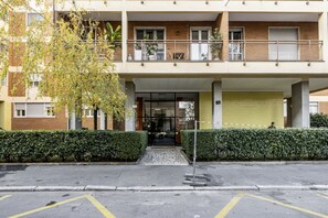 Un immeuble à plusieurs étages, doté d’une entrée fermée par un portail, d’un espace couvert avec un volet jaune et d’un arbre au premier plan.