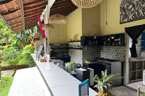 Egy konyha, amelyben egy hosszú konyhapult, különféle háztartási gépek találhatók, és egy kis kert is tartozik hozzá, növényekkel és egy sétánnyal.